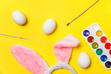Happy Easter concept. Preparation for holiday. Decorative eggs colorful paints and bunny ears furry costume toy isolated on trendy yellow background. Simple minimalism flat lay top view copy space