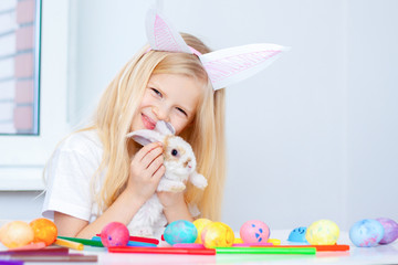 Blonde girl in rabbit ears on head and little bunny in her hands. Colorful eggs and markers on table. Prepearing for Easter and holidays concept