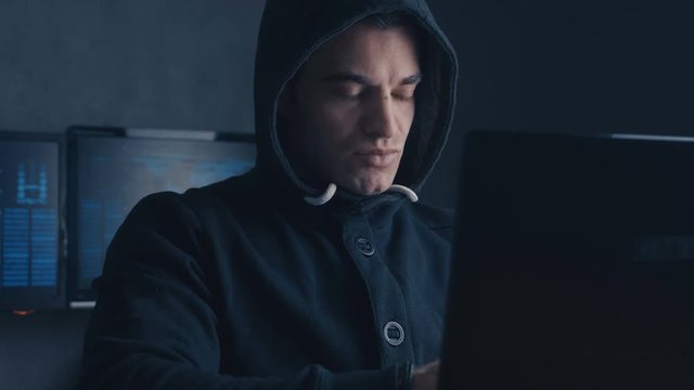 Cybercrime concept. Wanted Man hacker in hood working on a computer in a dark office room