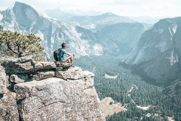Young man sitting on the very edge of the cliff admiring Yosemite National park half dome cliff