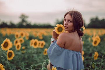 Dreaming young woman in blue dress holding and sniffing a sunflower in a field of sunflowers at summer, view from her back. Looking back