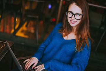 Woman in  the blue sweater uses a laptop at the workplace and holding a cup of fruit tea in hands, working remotely at cafe. 