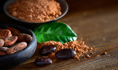 Cocoa beans and powder in bowl with leaves on wooden table