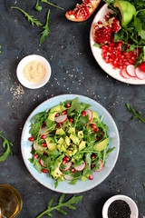 Ingredient for making salad on dark background. Vegetable salad in plate, avocado, arugula, pomegranate, radish and sesame. Top view with copy space. Vegetarian and vegan food.
