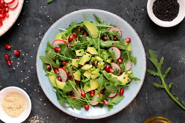 Ingredient for making salad on dark background. Vegetable salad in plate, avocado, arugula, pomegranate, radish and sesame. Top view with copy space. Vegetarian and vegan food.