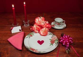 Floral pattern fine china dinnerware with matching plate, cup and saucer. bouquet of orange and white roses, pink napkin, silverware, red candles and card