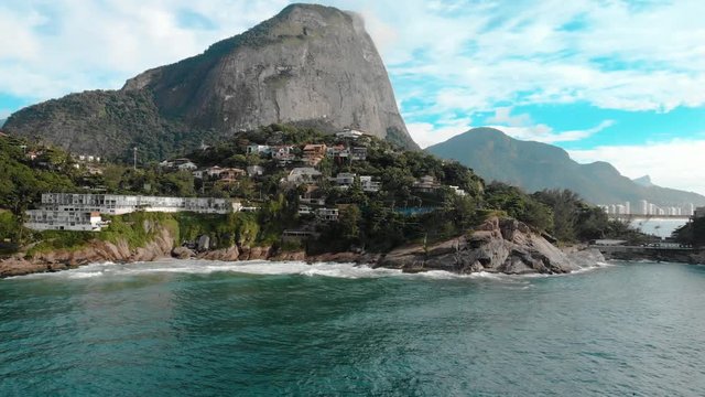 Aerial panning up showing the beautiful picturesque cliff rocks of Joatinga beach in Rio de Janeiro with the Gavea mountain towering behind it and the green ocean waves rolling in
