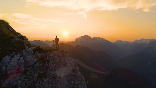 AERIAL, SILHOUETTE, LENS FLARE: Stunning sunset illuminates the Alps and hiker standing on edge of a cliff and observing the picturesque mountain landscape. Man watching the sunrise in the mountains.