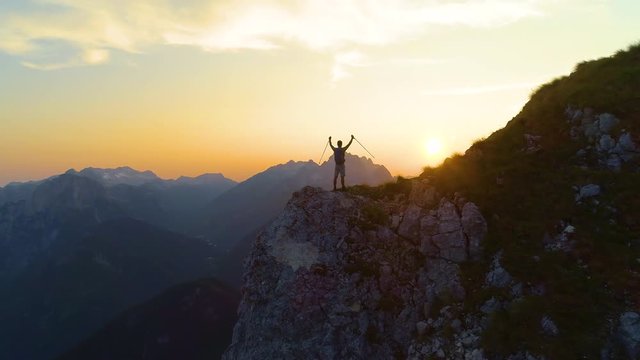 SILHOUETTE, DRONE, LENS FLARE: Golden morning sun rays shine on the ecstatic man hiking in the picturesque Slovenian Alps. Young male hiker celebrates reaching the mountaintop to see the sunrise.