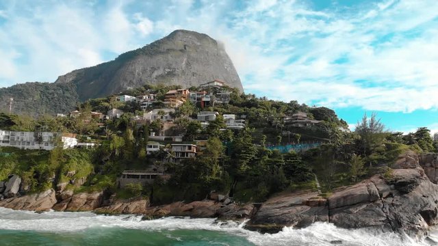 Aerial distancing of the beautiful picturesque cliff rocks of Joatinga beach in Rio de Janeiro with the Gavea mountain towering behind it and the green ocean waves rolling in
