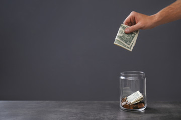 Man putting money into donation jar on table against grey background, closeup. Space for text