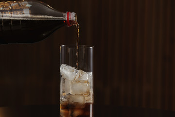 Pouring cola from bottle into glass with ice cubes on table against blurred background. Space for text