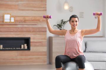 Fototapeta na wymiar Young woman doing exercise with dumbbells on fitness ball at home. Space for text