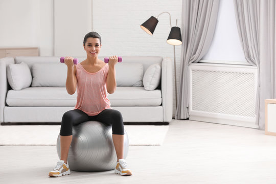 Young woman doing exercise with dumbbells on fitness ball at home. Space for text