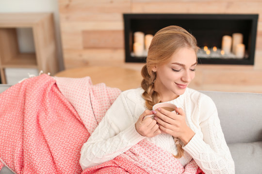 Beautiful young woman wrapped in plaid sitting on sofa with cup at home. Winter atmosphere