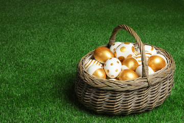Wicker basket of traditional Easter eggs decorated with golden paint on green lawn, space for text