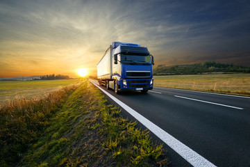 Blue truck arriving on the asphalt road in rural landscape at sunset