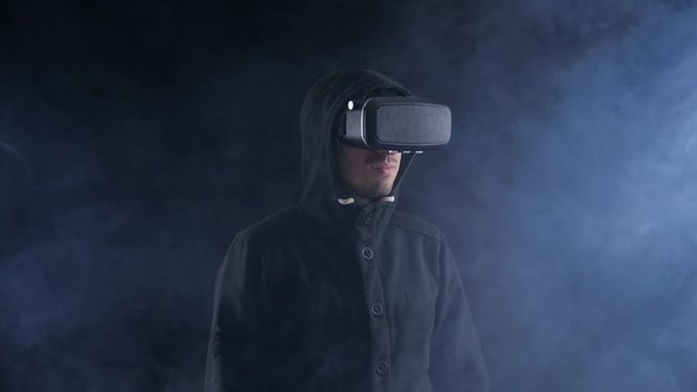 Futuristic man in hood Wearing VR Headset. Futuristic man using Virtual Reality Glasses in a dark smoky room.