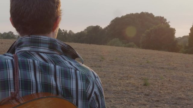 A lost young man with guitar watches the sunset on golden sunny field