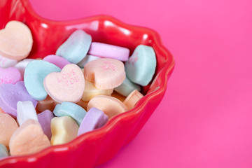 Colorful conversation heart candies in a heart shaped, red bowl against pink background. Valentine's Day theme.