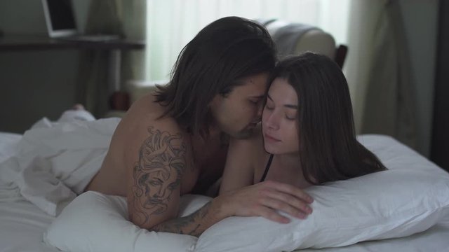 Handsome man cuddles his girl lying in bed and kissing her tenderly. Man caress his partner. Romantic relationship of beutiful couple. Lovers have a fun in comfortable room