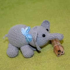 A crocheted gray elephant with a blue bow on the neck stands next to a bunch of wooden pencils on a green terry background.