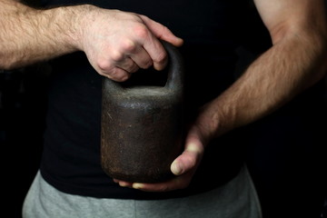 A man in a black T-shirt and sweatpants brutally holds a vintage weight in strong hands as a symbol of sport, strength, masculinity, fitness