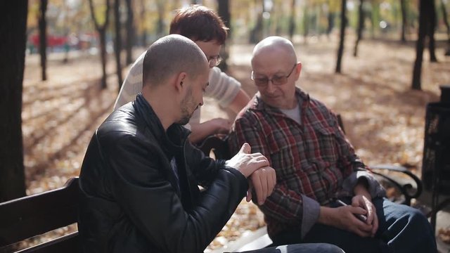 Elderly parents with adult son communicate in the autumn Park on a bench. The elderly man explains to parents how to use smart watch.