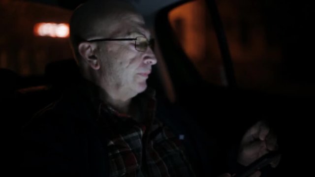 An elderly man using cell phone in a taxi. He is texting, checking mails, chats or the news online. Night.