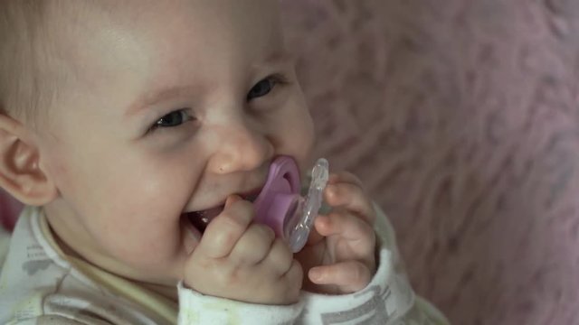 Close-up of baby girl's face with big blue eyes sitting in a crib smiling, laughing and nibbling a teether for teeth