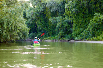 Woman paddle green kayak. Kayaking near overgrown shore of green thick thickets, wild grapes and driftwood on the banks of river at summer. Concept for adventure, travel, action, lifestyle.