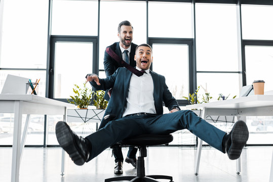 happy businessman pushing chair with excited coworker in modern office