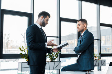 handsome businessman holding notebook and talking with colleague in modern office