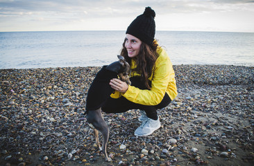 Young and brunette woman with her little dog on the beach with raincoat and hat.