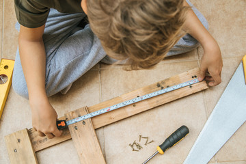Blond child sitting cross-legged on the floor is working with carpenter's tools measuring a wooden...