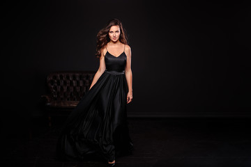 Portrait of young woman in elegant silk black dress posing in a dark interior, fashion beauty photo