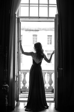 Black white portrait of young woman in elegant silk black dress lookingto a city from a balcony, fashion beauty photo
