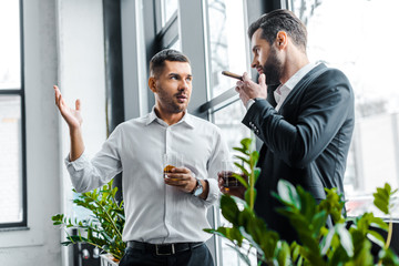 businessman holding glass of whiskey and having discussion with coworker with cuban cigar in hand