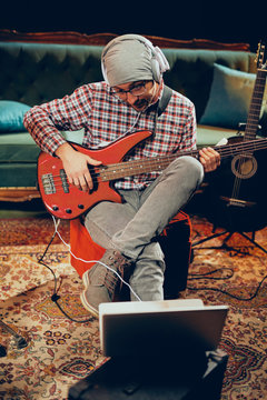 Young Caucasian man with hat on head playing bass guitar while sitting in home studio.