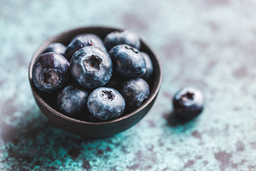 Fresh ripe blueberry in a small black bowl on a textured blue table. Macro photography.