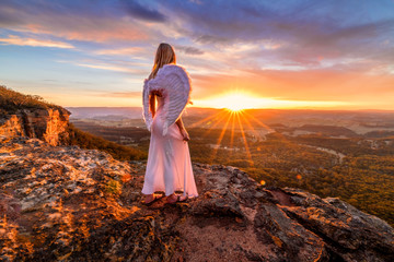 Angelic woman with angel wings and white dress on mountain cliffs sunset