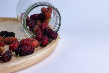 Mulberry fruit mulberry many both purple red orange in jar glass and mulberry outside jar in the plate made of wood all laid are on a white background.