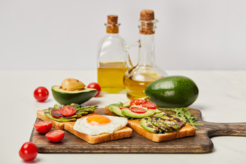 selective focus of wooden cutting board with toasts, scrambled egg, cherry tomatoes, avocados and oil bottles on grey background