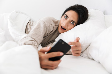 Photo of adorable woman 30s using smartphone, while lying in bed with white linen at home