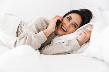 Image of smiling woman 30s using smartphone, while lying in bed with white linen at home