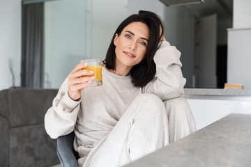 Image of caucasian woman 30s drinking orange juice, while resting in bright modern house