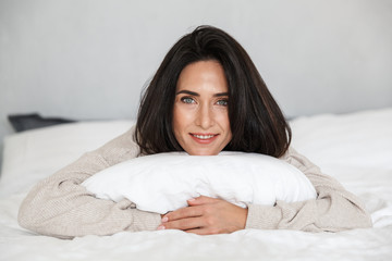 Photo of middle-aged woman 30s smiling, while lying in bed with white linen at home