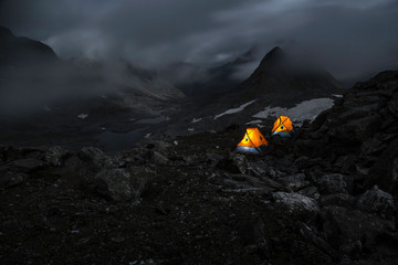 Night mountain landscape with two oranges illuminated tents. Silhouettes of mountain peaks with clouds. 