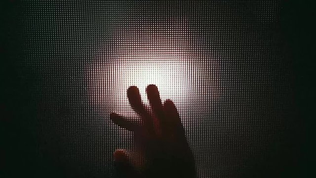 The silhouette of a woman's hand on glass emerges unexpectedly from the dark against the background of a bright searchlight. Patterned, tenxtured glass in the form of squares. The concept of horror.