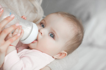 Young mother feeding her little cute baby daughter with bottle of child formula. Woman with her newborn baby at home. Mom taking care of a child. Alternative to breast feeding. - obrazy, fototapety, plakaty